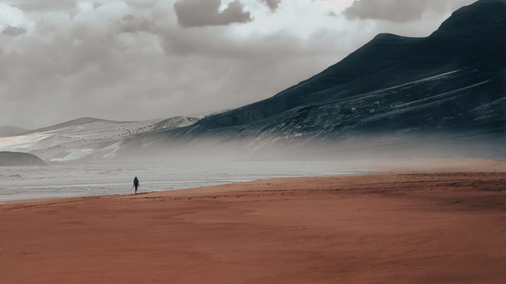 una persona passeggia in riva al mare, di fronte ci sono nebbia e montagne